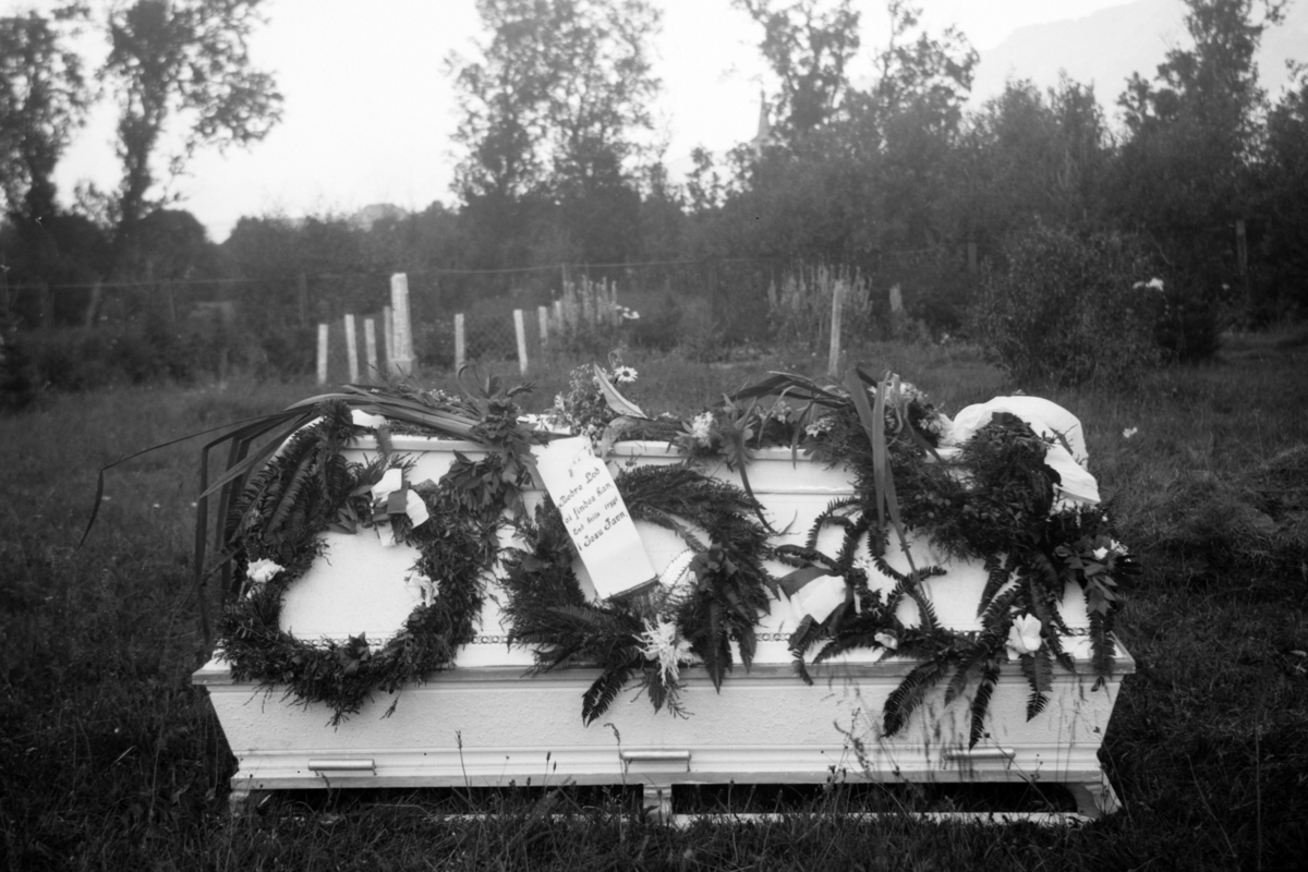 Hvit kiste, pyntet med blomsterkranser, fotografert utendørs. Bildet er trolig fra en begravelse i Ibestad.