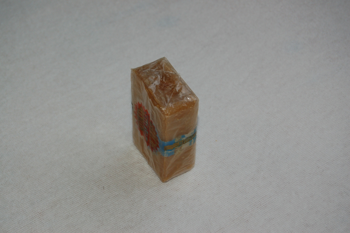 Rektangulært såpestykke innpakket i tynn plastfolie med trykt etikett. Tekst innprega i såpestykket, skjult av etikett.