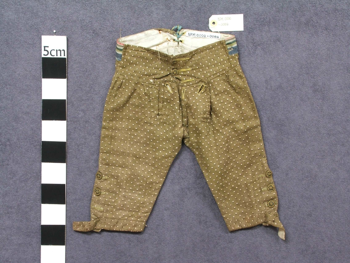 Jakke og bukse i bomullsstoff. Til gut. Høyrer saman som ei drakt.  Fôra med ufarga lin. Gul tråd i saum. Knappar i gull og svart. Buksa har klaff framme med tre knappar. Tre knappar nedst på kvart bein. Heilt nedst klaff med knapphol, her er knappane vekke. To knappar i lininga.  Knyting bak. Ein lapp i kvar side av lining. Jakka har silkeslag med påsydd pynt. Fire knapphol, berre ein knapp att. Fire hektar i heimelaga kvalitet til å hekta saman jakka øvst. På kvar side på jakka er det sydd på ein knapp med stoff.