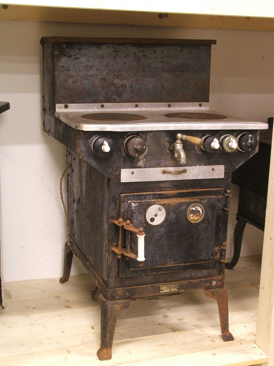 Flølo komfyren. Elektrisk komfyr. Med tank for varmt vatten (oppå bak), og kran for tapping av det varme vatnet framme. To kokeplater oppå. Fem runde brytarar framme i front. Ei uttrekkbar plate under. Omnsdør til å opne inn til steikjeomnen under der att. To plater med handtak inne i steikjeomnen. Komfyren står på fire bein (av jarn).