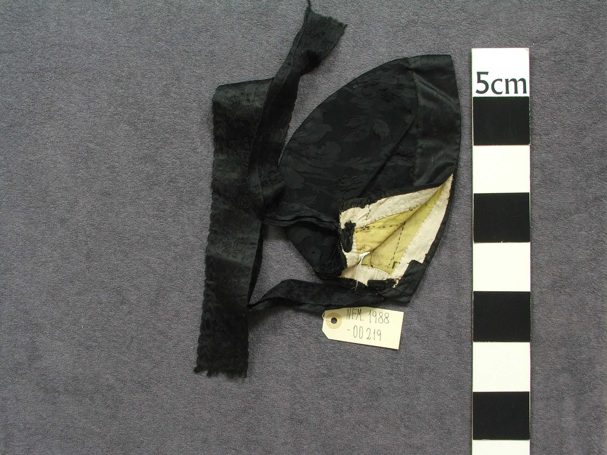 Svart "hue" til kvinne. Konehue. Typen er tett inntil hovudet. Med 4 cm kant fremst i glatt stoff, og med resten av utsida i svart stoff med innvove blomemønster.Lange band bak i nakken til knyting. Bak i nakken ein om lag 11 cm høg splitt, øvst i splitten er bandet trædd inn og i to hol på kvar side. Det er altså eitt band som kan snurpast ihop her. Fôra med lys gult og kvitt bomullsstoff.