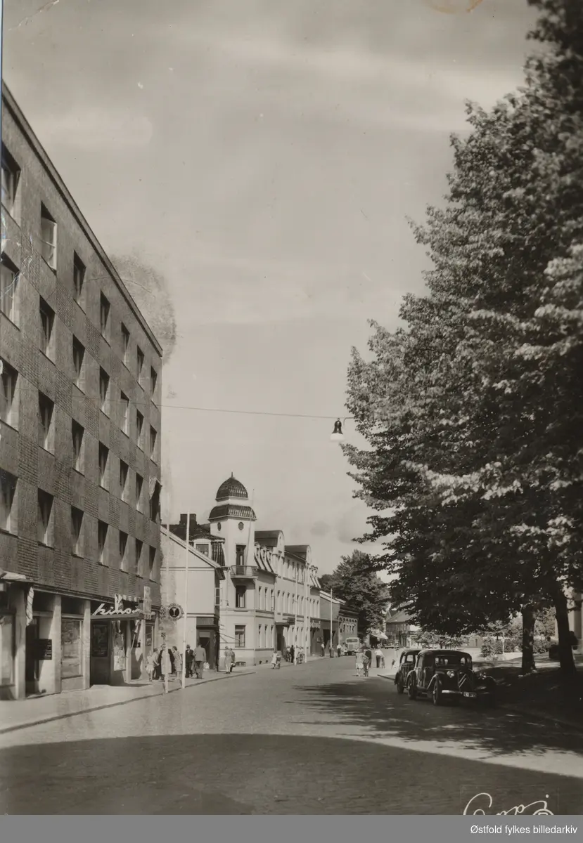 Dronningens gate  i Moss med kino til venstre glassmagasin, Moss hotell og Sparebanken. Til høyre står to biler parkert mot Kirkeparken. Foran en Citroên. Tittel på postkortet:"Moss. Dronningens gate"