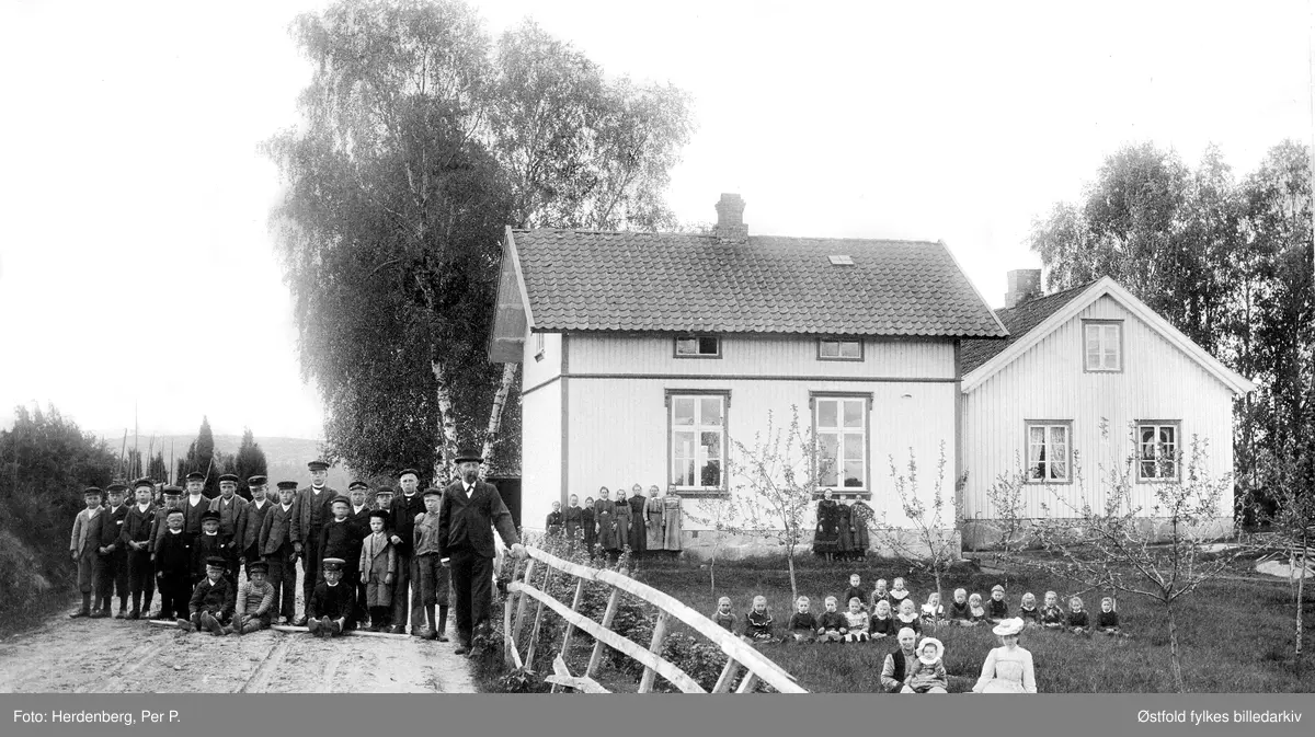 Nylende skole i Rolvsøy på slutten av 1890-tallet. Elevene er plassert i grupper; guttene til venstre på veien, de store jentene ved ny- bygget, de mindre jentene sitter i haven, og foran er lærerens familie. Lærerboligen ble skole og nybygg til lærer ferdig i 1892. Lærer Josef Olsen Hestenæs F.1853, kona Oline også lærer.