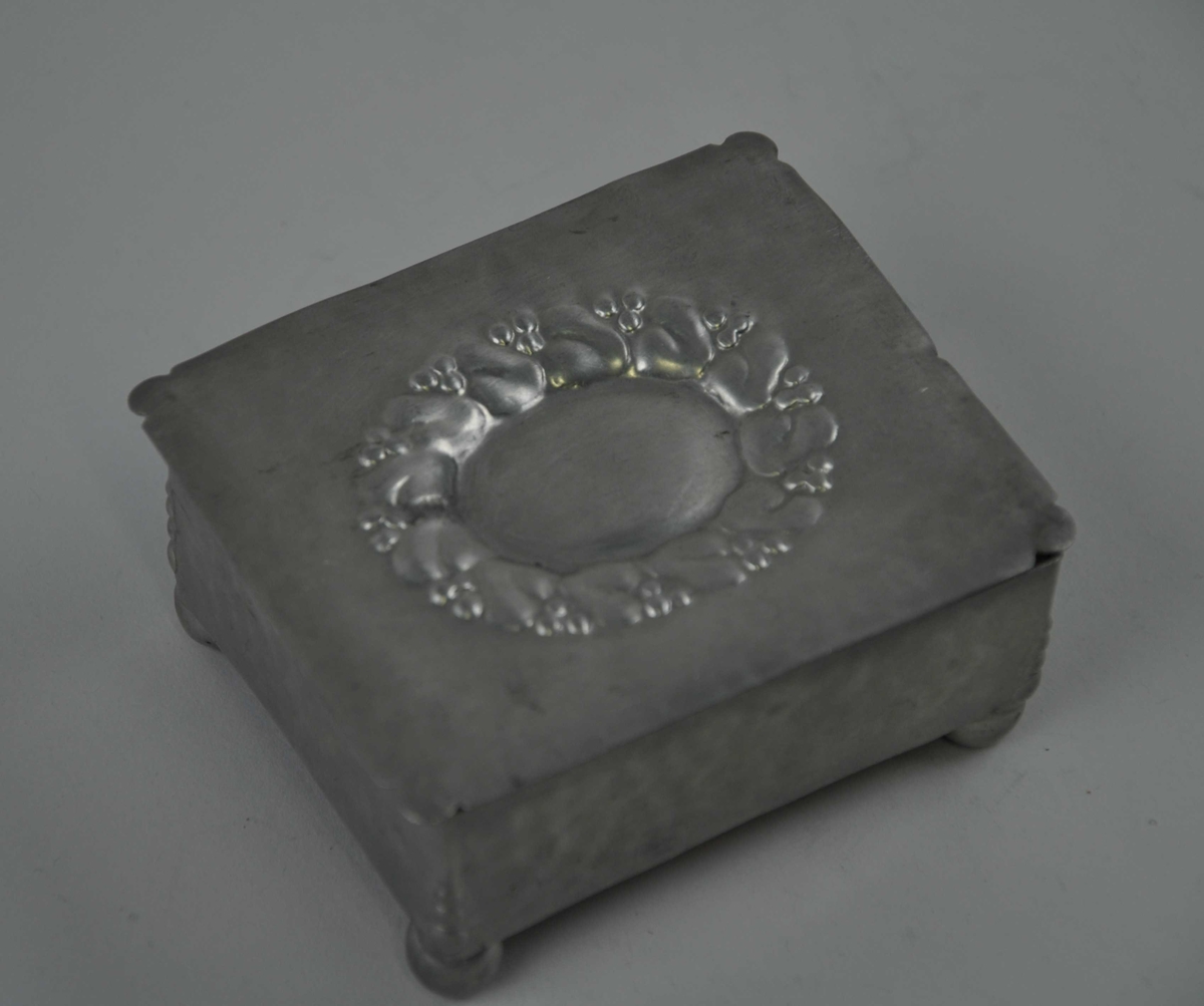 Oval  jugendstilornamentikk på loket i form av stiliseerte blad med bær. Bæra går att i hjørnedekorensom endar som kuleforma føter.