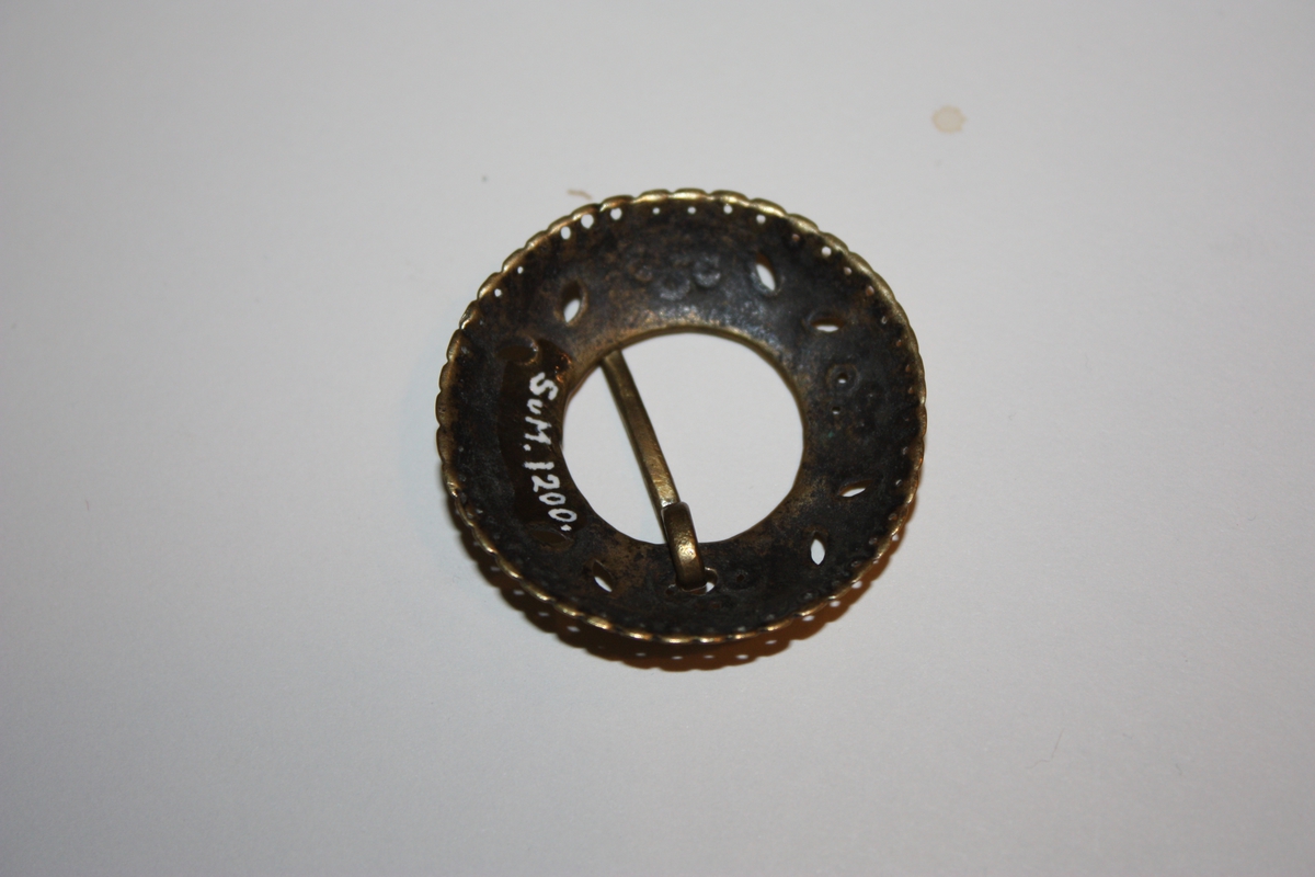 Ringforma med gjennombroten dekor og bølgja kant med hol innanfor. Gravert. Torn på oversida. Nytta som skjorteknapp