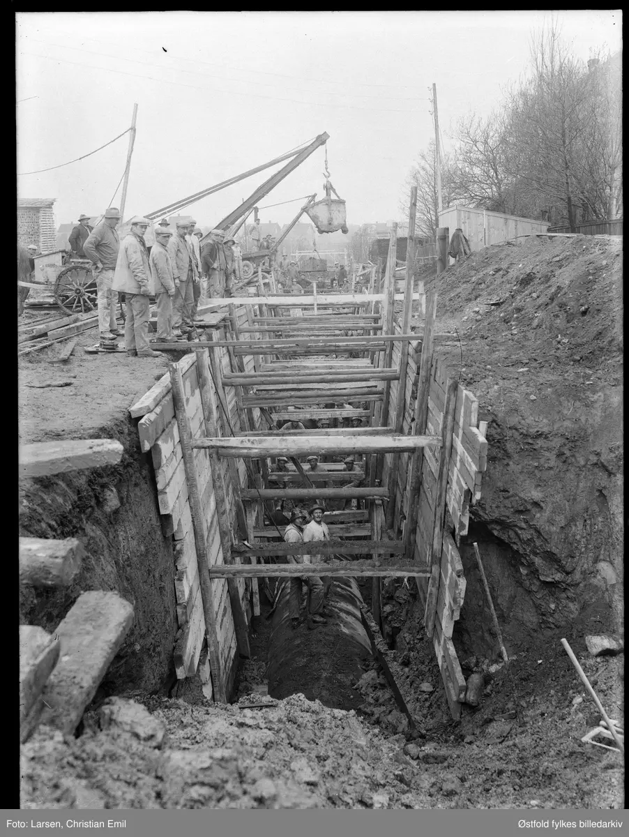 Graving av dyp grøft med forskaling, rør i bunnen, kjerre, heisekran, mange arbeidere i sving. Ukjent sted i Sarpsborg. Er årstall 1942 feil? Ser eldre ut.