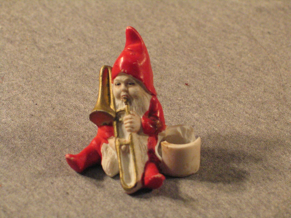 Nisse som sit og spelar trekkbasun. Ved sida av har han ei bøtte som det kan settast juletrelys i. Nissen har langt skjegg og hue med spiss.