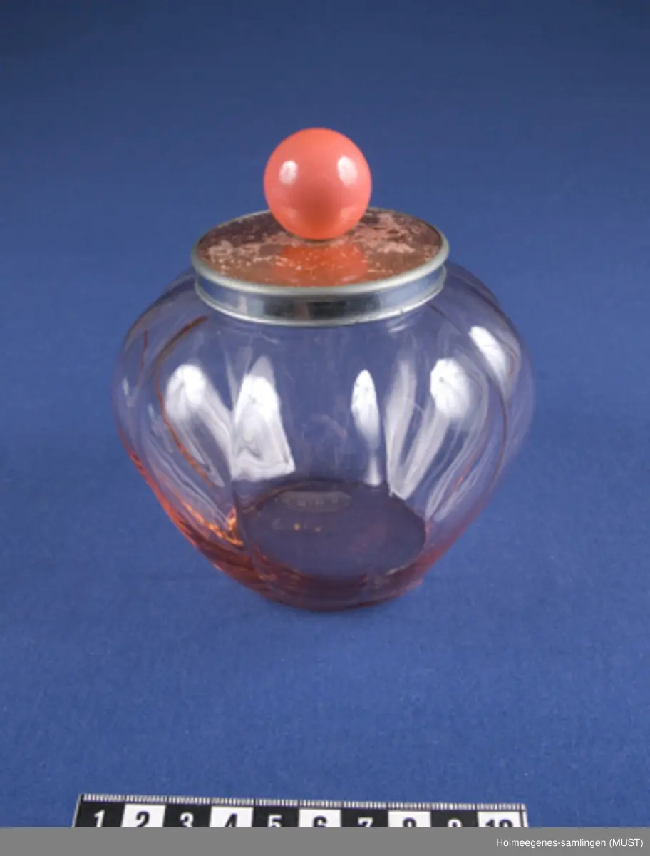 Bolle av formpresset rosa glass. Lokk av blikk med guillochering, belagt med rosa plast med knott av plastlignende materiale. (Guillochering er en maskinell metode å behandle sølv eller lignende materiale for å gi den et regelmessig rillemønster eller et geometrisk mønster som skimrer gjennom emaljen, her plasten.
