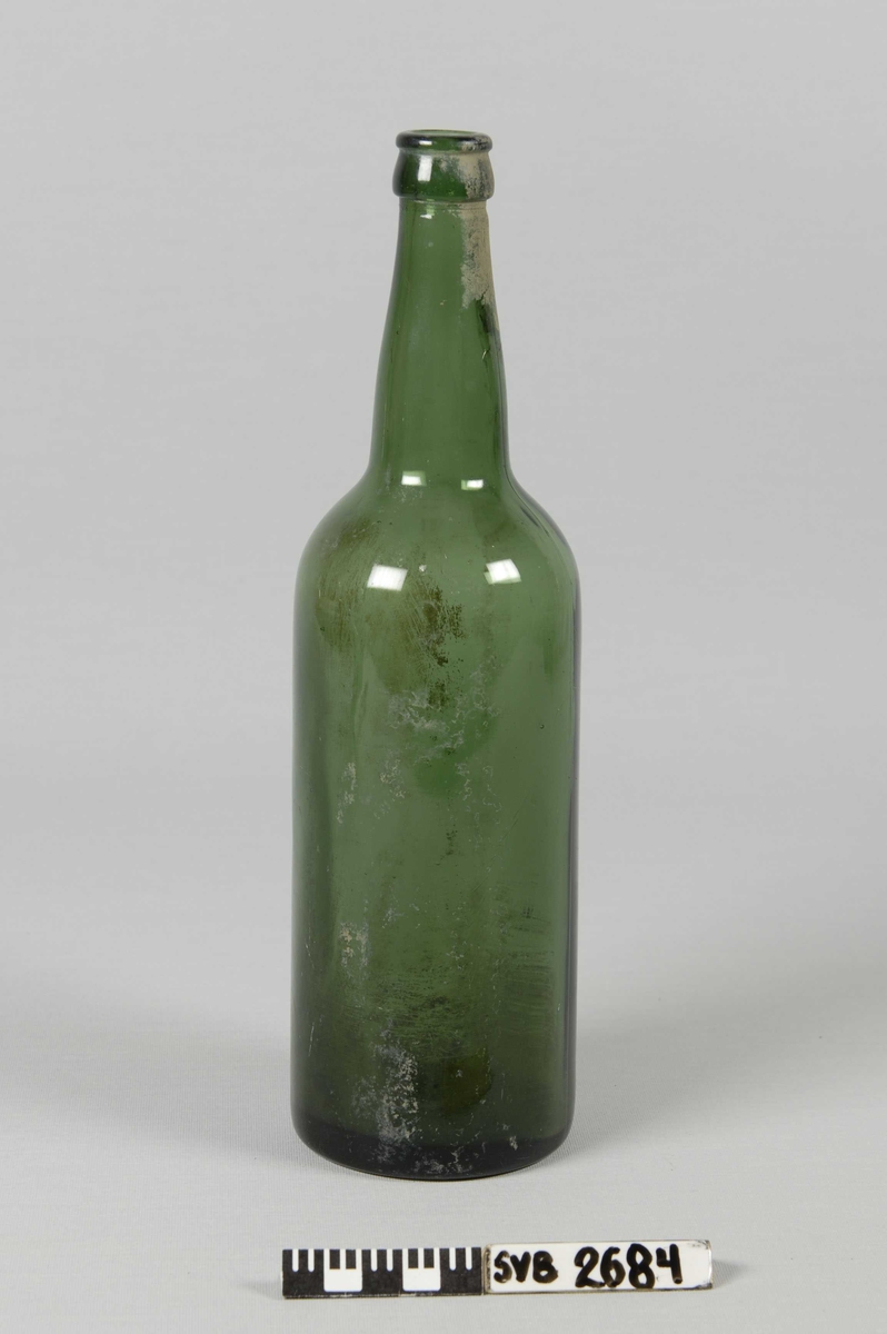 Drikkeflaske i grønt, klart glass. Merket "M" på flaskebunnen. En del leire på flasken. 