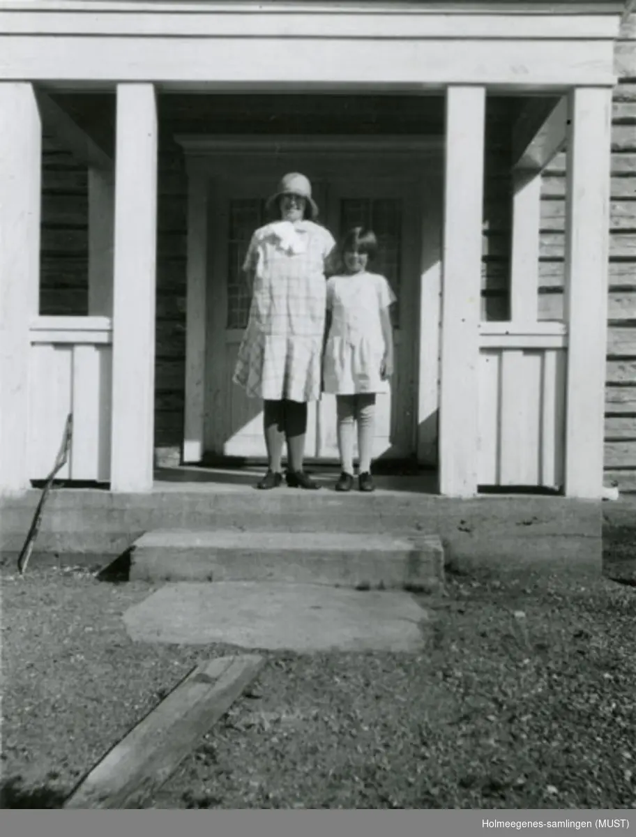 To jenter står på en overbygd trapp på et tømret hus. Antatt samme hus som ST.K.HE 2007-011-0120. Se også ST.K.HE 2007-011-0111 til -0126.