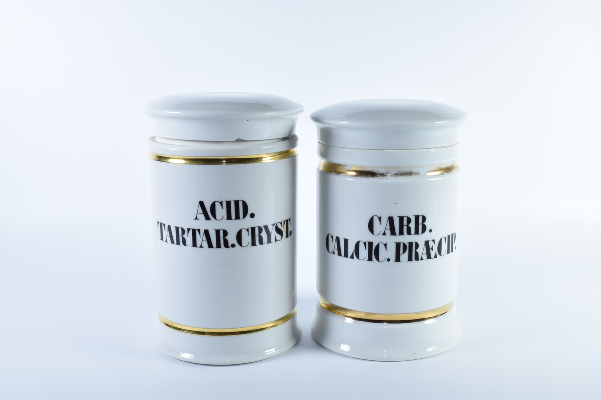 Seks sylindriske porselenskrukker med flatt lokk. Samtlige er dekorert med gullborder øverst og nederst og har skrift i svart eller rødt.