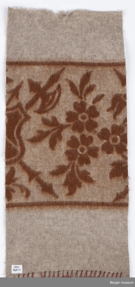 1 prøve. Teppet er ensfarget med en bred bord. I borden er det blomster og et våpenskjold med pil og øks.  Art Nouveau.