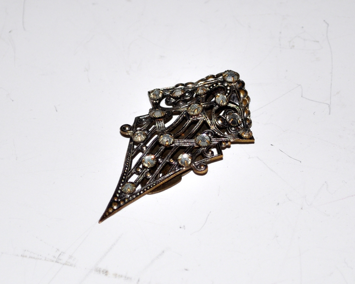 Fra protokollen: Nål, metall, 15 slepne "stener" (glass ant.) Støpt, imitert filigransarbeid. Gjennombrudt mønster. Clips til å feste nålen med på baksiden.