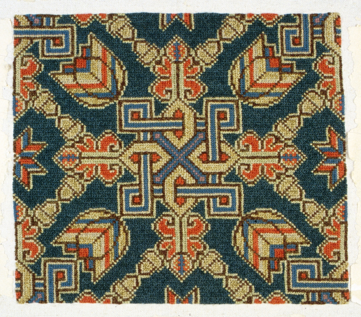 Tvistsömsbroderi troligen tänkt som framstycke till dyna/kudde. Broderiet kan också vara ett prov. Centrerat motiv med valknut, tulpaner och akantusblad samt diagonaler av ekollon. Broderad i två nyanser blått samt rött, grönt, gulgrönt, brunt, naturgrått och rosa. Sydd på panamaväv i bomull, varp och inslag i 2-trådigt bomullsgarn, 14 x 14 trådar/cm, med tvåtrådigt ullgarn.Rester av fastklistrat papper i kanterna på bottentyget (från äldra montering). Bottentyget är 30 x 50 mm större än broderiet. Kastade kanter.