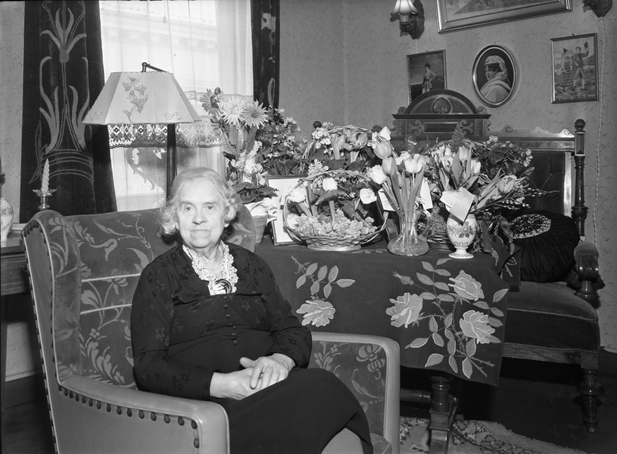 Caspara Nyblin f. Larsen sitter i en stol ved siden av blosmter. Åremålsdag med blomsterhilsener.