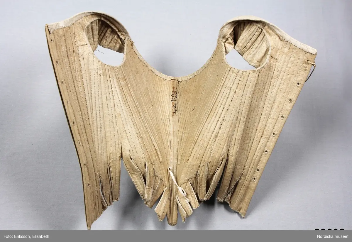 Huvudliggaren:
"Snörlif. Af hvitt siden, från 1770-talet."

Liv till hovdräkt s.k. "robe á la cour" av ursprungligen vit sidendroguet. Tyget förmodligen svensk tillverkning (Myrman?) se 1700-tals textil, A. Berchs samling, sid 171. Yttertyget är skuret i nio delar med mittstycke fram som löper ned i snibb samt fyra delar å varje sida med snörning mitt bak. Grundlivet tillverkat av oblekt linnelärft är helt styvat med spröt av valben trädda i sydda vertikala kanaler samt med en skena av järn som löper runt urringningen och planschett mitt fram. Grundlivet troligen klistrat med papper för att hålla en viss form innan yttertyget monterats på. Ärmholkar bestående av å varje sida tio hopsydda delar som lagts i veck. Tillskärningen följer helt den gängse för robe de cour. Se Waugh, Nora, The Cut of Womens Clothes, s. Längs sidorna i midjehöjd en tunn snodd avsedd att haka fast släpet i. Livet ursprungligen buret med kjol och släp i samma tyg.
/Ingrid Roos 2007-03-06