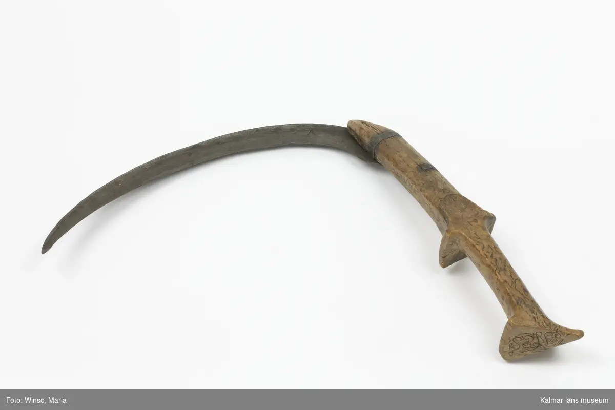 KLM 3120. Skära, vinkelskära, trä, metall. Vinkelskära med bågböjt blad av järn, fastnitat på skaftets ena sida. Skaft av masurbjörk med fyrsidig knopp med skarp mittrygg. På knoppens översida inskuret årtalet MDCCCI (1801 och blommor och rankor). På handtaget nära bladet inbränt PNS. Bladet märkt A. Användes för att skära av säden.