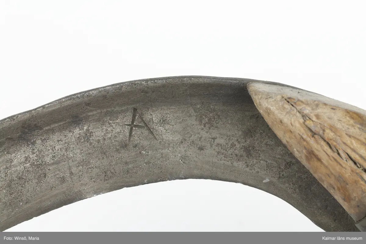 KLM 3120. Skära, vinkelskära, trä, metall. Vinkelskära med bågböjt blad av järn, fastnitat på skaftets ena sida. Skaft av masurbjörk med fyrsidig knopp med skarp mittrygg. På knoppens översida inskuret årtalet MDCCCI (1801 och blommor och rankor). På handtaget nära bladet inbränt PNS. Bladet märkt A. Användes för att skära av säden.