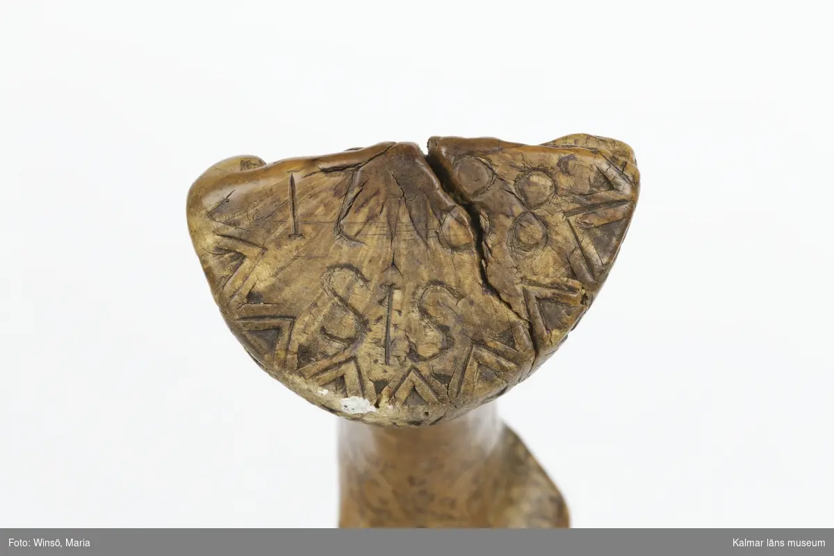 KLM 7163. Skära, vinkelskära. Av trä och metall. Vinkelskära med bågböjt blad av järn, fastnitat i skaftet. Skaft av masurbjörk, ornerat med plattsnidat rutmönster. Ornerat vågmönster på bladets ena sida samt inpunsat: ANO 1788 SIS. Rund knopp, en bit saknas. På knoppen skuret sicksackmönster samt 1788 SIS. Användes för att skära av säden.