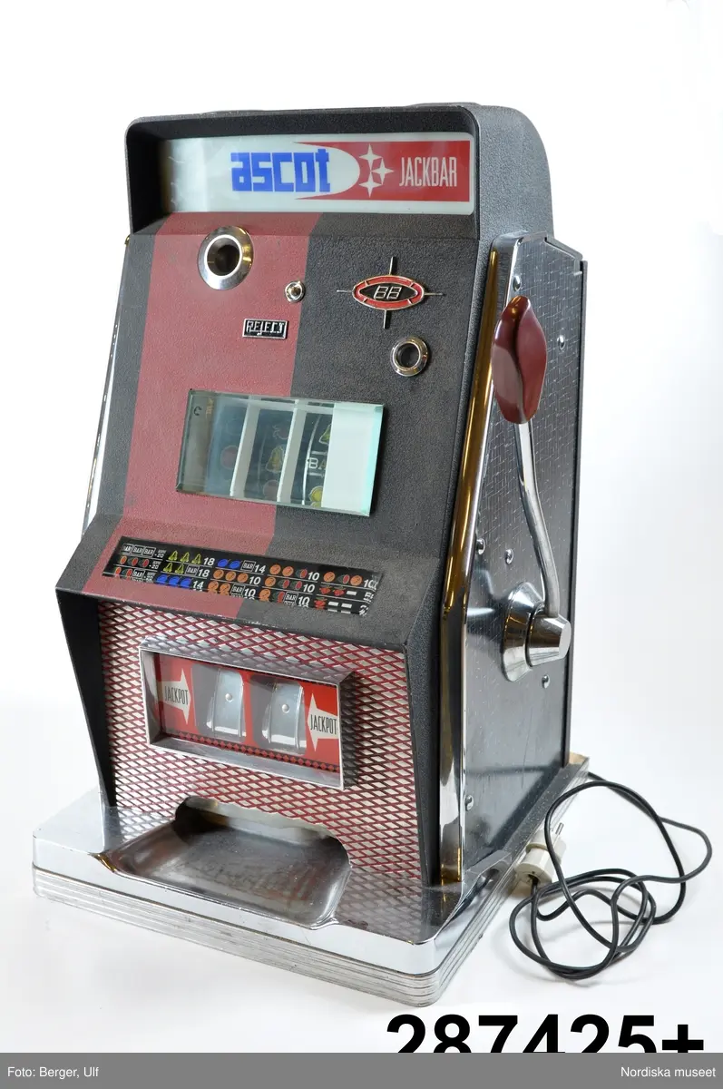 Inventering Sesam 1996-1999:
L 46 cm
B 44,5 cm
H 74 cm
Spelautomat, enarmad bandit. Hölje av svart och röd metall och svartvit perstorpsplatta. Stående på träplatta med vitmetall. Framtill olika skyltar bakom tjockt glas. Mitt på (eg. innuti) tre rullar med olika symboler, t ex apelsiner, halva meloner, siffror mm, bakom glas. På vänster sida hål för mynt. På höger sida en arm med rött handtag av backelit. På vänster sida elektrisk sladd med vit kontakt. 
Text på skyltarna text "ASCOTT JACKPOTT", "Reject", Jackpott" och karta med de olika symbolkombinationer som man vinner på.
Innuti mekanism av metall. Öppningsbart bakstycke (med nyckel vilken saknas vid inv. 1998) 
Liten svart skylt nertill på vänster sida; "Ascott s.p.a. / Calenzo - Florence Italy / Tipo 88 / M.la 067"
Funktion; man stoppar in ett mynt (troligen 1 krona) och drar i armen. Rullarna med de olika symbolerna surrar och stannar sedan en och en (går ev att själv stoppa med en liten knapp mitt på framsidan). Om man lyckas få någon av de kombinationer som finns på skylten så vinner man pengar, som kommer ut längst ned. Mest vinner man om man får tre lika symboler. Enarmade banditer har varit vanliga på motell, konditorier, bensinstationer och dyl. 1973 fanns ca 140 000 spelautomater, samma år infördes skatt på automaterna. Senare blev de förbjudna. 
Jmfr liknande enarmad bandit av samma tillverkare (?) inv.nr 287.426
Bilaga
Leif Wallin feb 1998