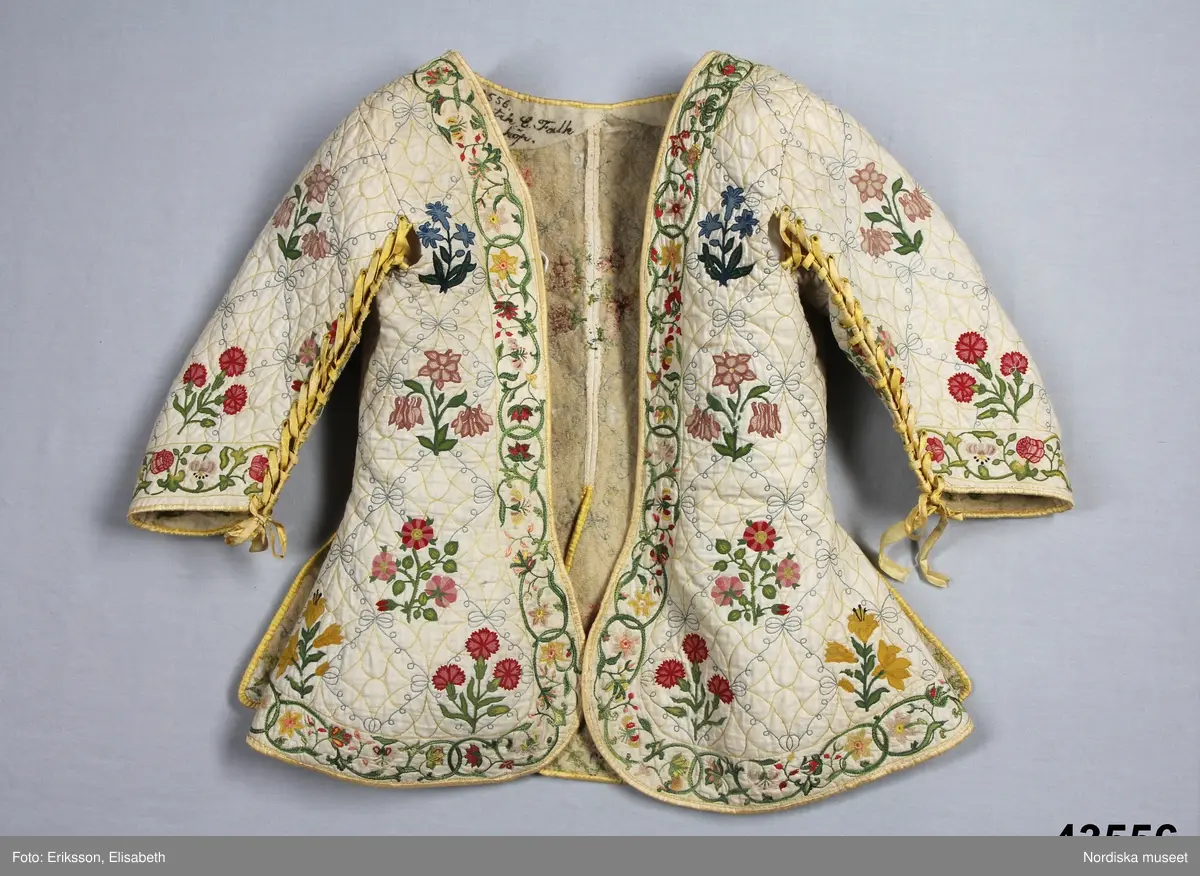 Katalogkort IV:
"Tröja / kofta, för dam, 1600-talets slut.
Längd 54 cm
Av linnelärt med foder av glest tuskaftat ylletyg. Försedd med stickningar i gult och blått samt broderier i kedje-söm med yllegarn. 2 ryggstycken, 2 framstycken. Halvlånga ärmar med 2 sömmar, varav den ena öppen och kantad med gult siden samt försedd med gult silke tränsade snörhål, med band av dubbelvikt sammansytt band. Sömarna uppslitsade nedifrån upptill midjan och kantade med gult siden."