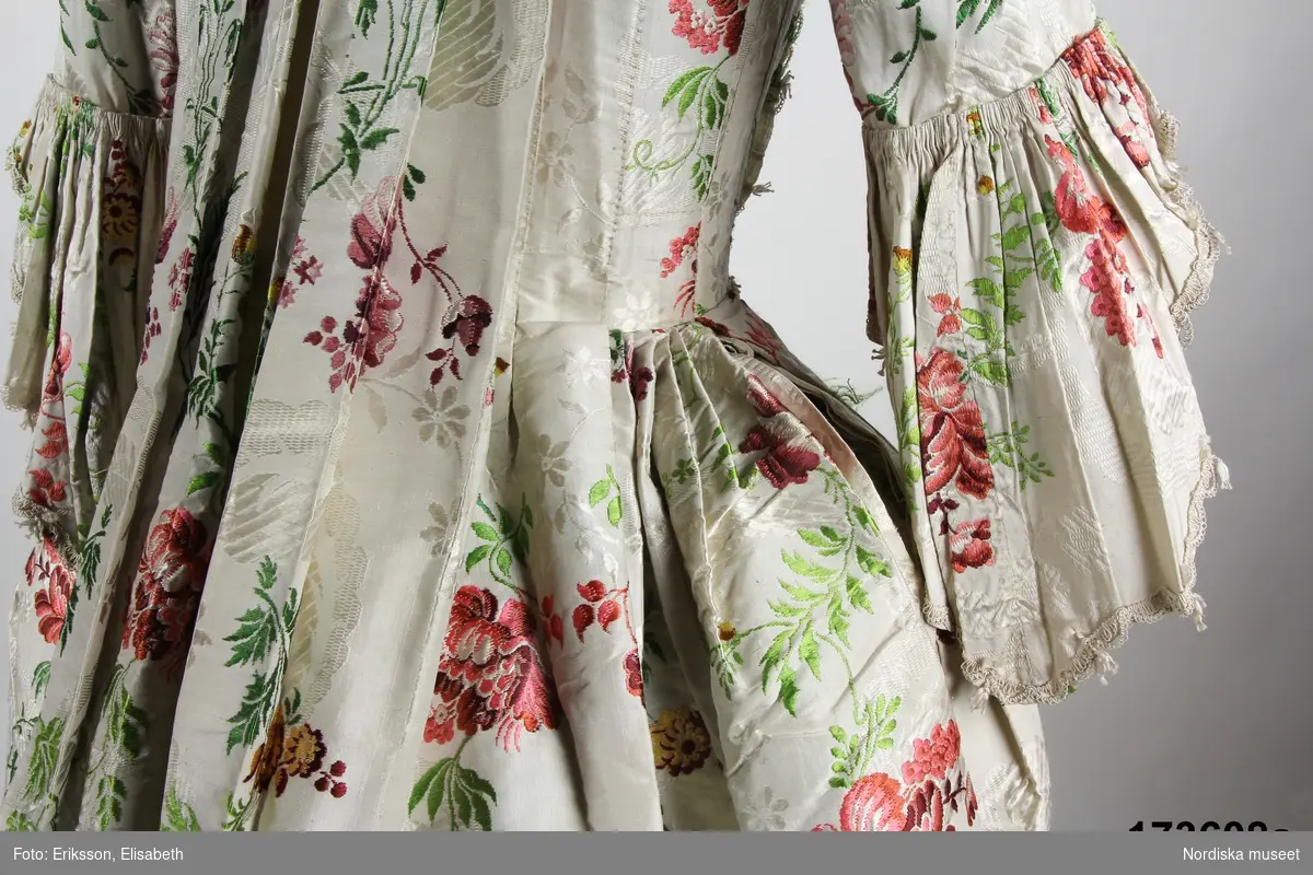 Äldre katalogkort: 
"Robe och kjol  av vit sidenbrokad med blommor i rött, gult m.m. Robe med dubbla watteauveck och engageanter av vita knypplade silkespetsar i mångdubbla rader. Livet fodrat med vitt linne med fiskben i sömmarna. Bröstlapp saknas.
Tygets vådbredd 50,5 cm. Tygåtgång ca 13,5 meter."

Bandgarnering av veckad remsa av tyget kantat med uddganser i vitt silke.
I utmärkt fint skick.
/Berit Eldvik 2009-01-09