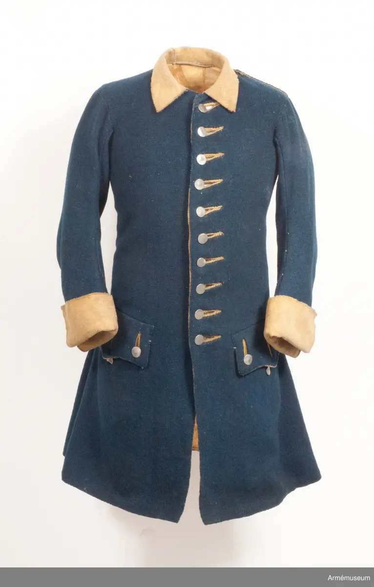 Rock av blått kläde med gul krage och ärmuppslag. Fodrad med gul boj i livet och med linne i ärmarna. Försedd med tennknappar.