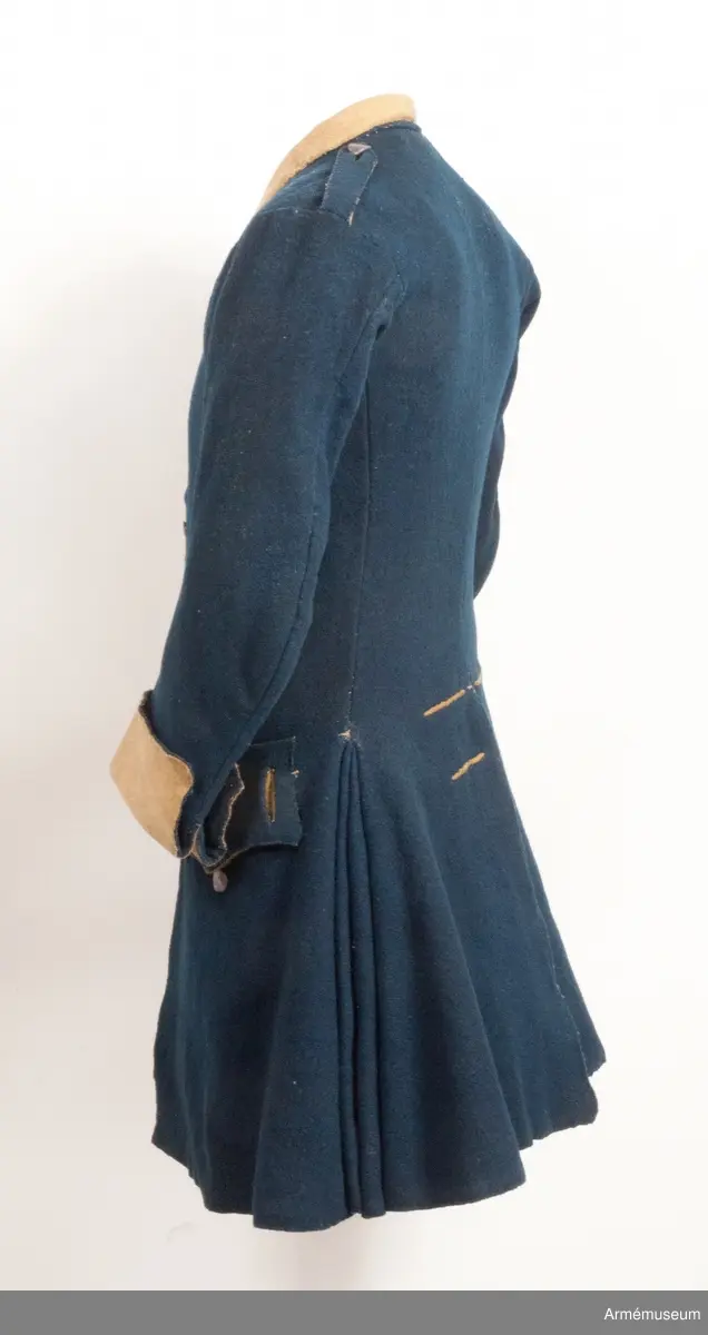 Rock av blått kläde med gul krage och ärmuppslag. Fodrad med gul boj i livet och med linne i ärmarna. Försedd med tennknappar.