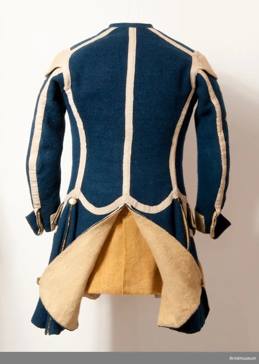 Rock med prydnader för spelet; mellanblå med gul rabatt (bröstrevärer), svalbon (axeldekoration) och ärmuppslag. Vit redgarnsdekor bruklig för spel. Rocken har sammanlagt 23 knappar. Knapparna i framkanten fastträdda med vitt band från avigsidan. En knapp i vardera skört (en ny). Två knappar på ficklocken (höger sida fattas en). Rocken är fodrad med gult ylletyg. Rocken ihopfästes med hyskar och hakar.