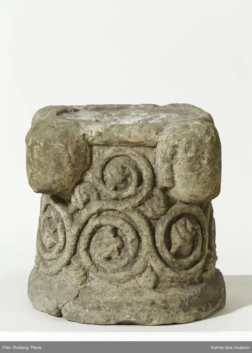 KLM 35314. Dopfunt, dopfuntsfot. Av kalksten. Fot till dopfunt med rund genom skärning. Spiralupprullade växtrankor i relief samt upptill fyra kraftigt utstående ansiktsmasker. Ett cylindrisk hål tvärs igenom stenen.