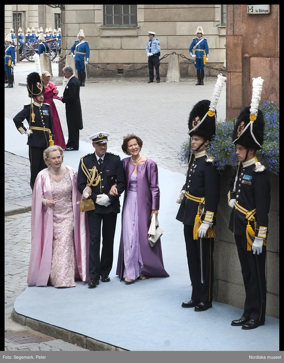 Kronprinsessbröllopet. Dokumentation av bröllopet mellan Kronprinsessan Victoria och herr Daniel Westling 19 juni 2010. Gästerna anländer till Storkyrkan. Marianne Bernadotte i ljusrosa taftklänning med metallsilverspets och aftoncape i taft, diadem och smycken. Madelene Kogevinas i ljuslila långklänning och mörklila aftoncape, diadem och smycken. Man i stor mässdräkt.