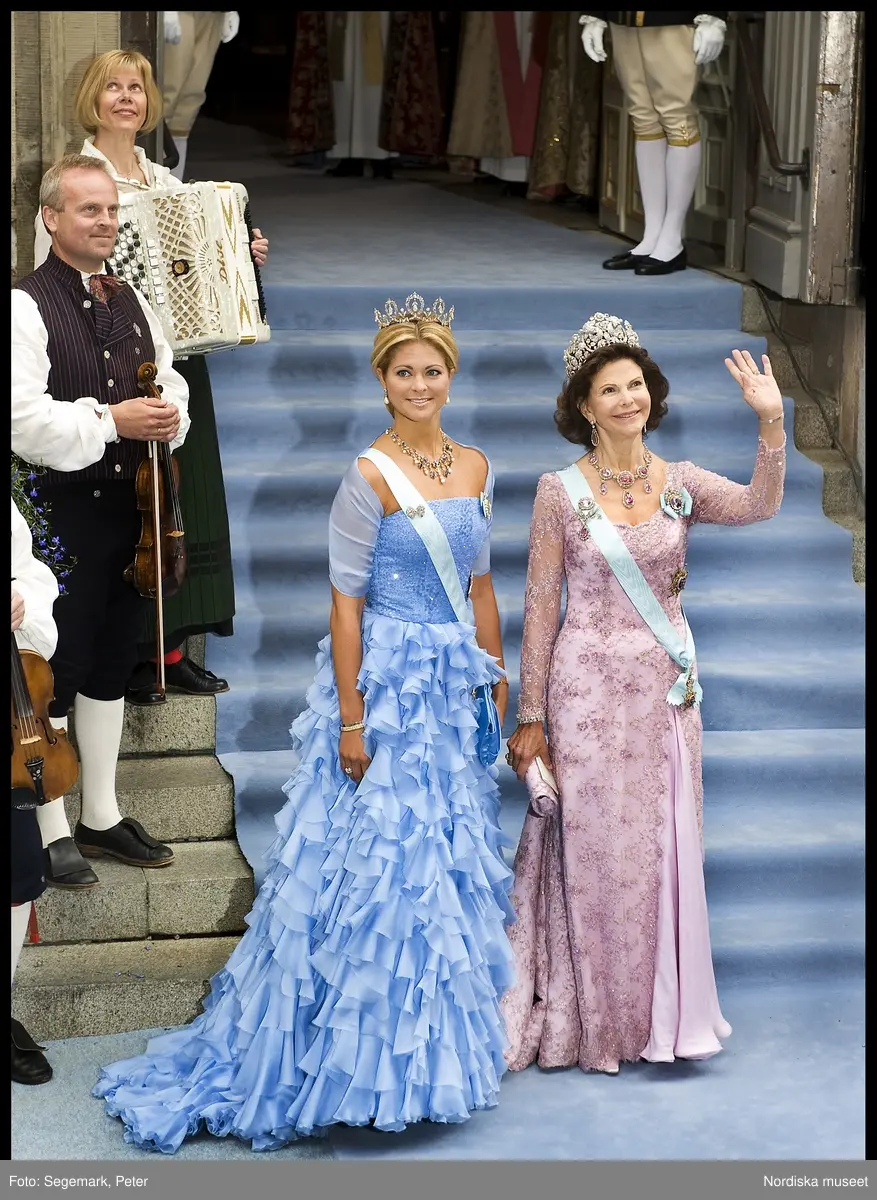 Kronprinsessbröllopet. Dokumentation av bröllopet mellan Kronprinsessan Victoria och herr Daniel Westling 19 juni 2010. Gästerna anländer till Storkyrkan. Drottning Silvia i rosa tyllklänning med pärl-och stenbroderier och silvrig calaisspets, det brasilianska diademet, halsband, örhängen och brosch av diamanter och rosa ädeltopaser, ordenstecken. Prinsessan Madeleine i förgätmigej-blå chiffongklänning med kristallbroderat liv, prinsessan Sibyllas diadem av fem blomsterkransar i briljanter och dallrande briljantkläppar, halsband-intågssmycket uppbyggt av ceylonsafirer, barockpärlor och briljanter, ordenstecken.