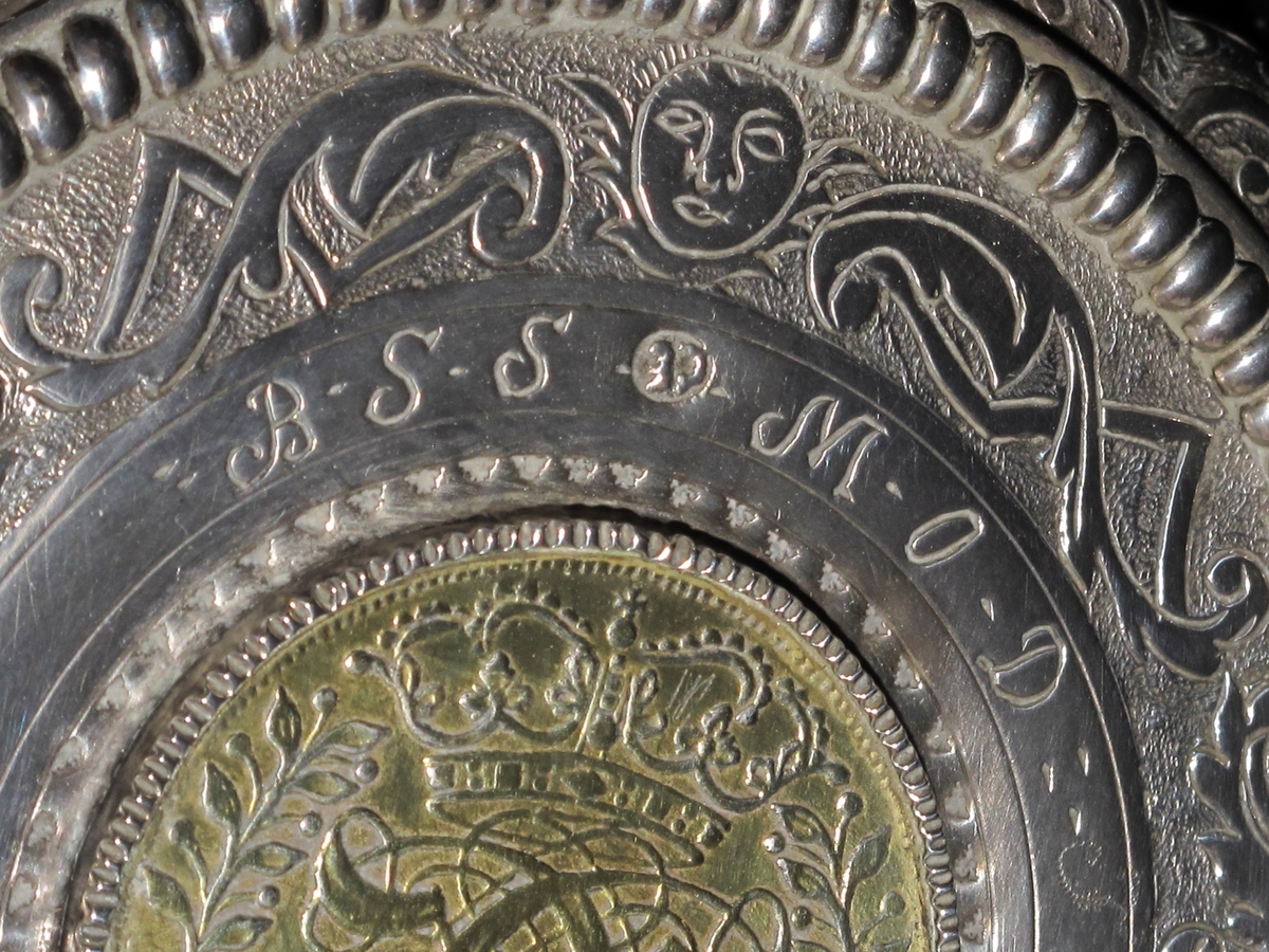 Stentøykrus m. sølvlokk. Lokket har en profilert, knekket kant m. regenceornamentikk, flat  toppflate, begrenset av opphøyet, tverriflet ring,  graverte blad og knekkete bånd på punslet bunn, i midten  mynt i smal riflet innf., som lokkgrep stående blad  m. opprullede fliker. Mynten C5´s kronede speilmonogram omgitt av kryssede bladgrener, reversen kronet riksvaaben og   IIII MARCK DANSKE 1694,  forgyllt. Rundt mynten gravert BSS, MOD, 1748, nyere innprikket FBW. Stpl. CE  i ovalt felt. 
Sølvlokket er montert på stentøykrus, som er sylindrisk m. svakt buede sider, lysbrun  glasur m. forsenket hvit ring øverst og nederst, samt hvit bunn,  munningsring og hank,  denne liten, flattrykkt øreformet.