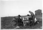 'Akka-expeditionen sommaren 1931: ::  :: 4 st män vid ett bord på en äng vid stranden. Leonard Axel Jägerskiöld andre från höger (sittande) med ryggen mot kameran. På avstånd en trälada. Se även fotonr. 2183. ::  :: Ingår i serie med fotonr. 2182-2234.'