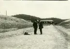 'Vy över fyndplatsen av Göteborgs Naturhistoriska museums subfossila kronhjortshorn.  Fynddatum: 1913-06-00. Med på bilden finns 2 män stående på en väg i tegelbruket. I bakgrunden skymtar en stor byggnad samt skog. ::  :: Se fotonr. 269:1-3. Se även fotonr. 620, 4478 och 1044.'