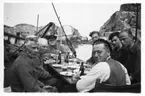 'Skandiaexpeditionen 1935: ::  :: 8 män kring dukat middagsbordet på Skandias däck i Moröns hamn. Längst bort på kortändan sitter Leonard Axel  Jägerskiöld. I bakgrunden berg. Främre kortändan Sune Swärd? ::  :: Ingår i serie med fotonr. 3441-3452.'