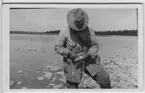 David Sjölander ringmärker skrattmås. Stående invid strand med hattklätt, nerböjt huvud. Blockrik strandkant. Vy ut över havet med barrskogsridå i fonden.