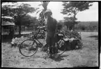 'Diverse fotografier från bl.a. dåvarande Nordrhodesia, nu Zambia, tagna av Konsul Magnus Leijer. ::  :: En man ståendes med en cykel, på grusväg/plan vid en rund blomsterplantering.'