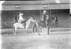 'Diverse fotografier från bl.a. dåvarande Nordrhodesia, nu Zambia, tagna av Konsul Magnus Leijer. ::  :: En man och en kvinna sittandes på en varsin häst. Fotograferade från sidan framför ett en tegelbyggnad, i en dörr skymtar ytterligare kvinna.'