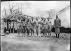 'Diverse fotografier från bl.a. dåvarande Nordrhodesia, nu Zambia, tagna av Konsul Magnus Leijer. ::  :: Åtta kvinnor stående på rad, bredvid dem står en man. Samtliga framför enklare byggnader/hyddor. Under ett halmtak sitter ytterligare tre peronser.'