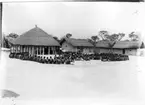 'Diverse fotografier från bl.a. dåvarande Nordrhodesia, nu Zambia, tagna av Konsul Magnus Leijer. ::  :: En stor grupp människor sittandes på marken framför en rund byggnad, på dess veranda sitter ytterligare människor, ett fåtal är stående.'