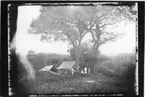 'Diverse fotografier från bl.a. dåvarande Nordrhodesia, nu Zambia, tagna av Konsul Magnus Leijer. ::  :: 1 st kvinna med 1 st barn sittade framför tält och träd, bredvid henne står 2 st män i uniform bakom står 5 st män. Placerade på öppen plats bland höga växter.'