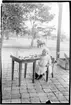 'Diverse fotografier från bl.a. dåvarande Nordrhodesia, nu Zambia, tagna av Konsul Magnus Leijer. ::  :: Litet barn sittande på en stol vid ett kaffedukat bord på en veranda. Bakom barnet är en fågel sittande på en låda samt en person vid en häst synliga.'