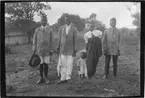 'Diverse fotografier från bl.a. dåvarande Nordrhodesia, nu Zambia, tagna av Konsul Magnus Leijer. ::  :: Tre män samt en kvinna med ett barn står på rad, i bakgrunden är ett staket samt träd och gräs synligt.'