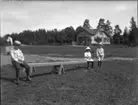 'Bildtext: ''Barn, Ed.'' :: Vy med öppen gräsmark och i bakgrunden ett hus. I förgrunden 3 barn som sitter på en bänk alternativt vippbräda. Lek. ::  :: Ingår i serie med fotonr. 5225:1-18. Se även fotonr. 5230:1-11.'