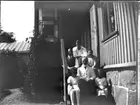 'Gruppbild, 1 man, 1 kvinna, 2 unga män/pojkar och 2 flickor sittande på trappa till hus. Klädsel. :: Ingår i serie med fotonr. 5226:1-11.'