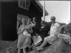 'Bildtext: ''Kyrkesund 27/7 1927.'' :: 3 kvinnor och 2 män sittande och stående på berghäll vid flaggstång, framför trähus. 1 av kvinnorna stickar, en håller en tidning och en av männen har en pipa i munnen. ::  :: Ingår i serie med fotonr. 5228:1-11.'