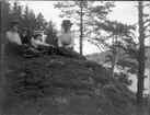 'Bildtext: ''Gruppbild Trolldalen.'' :: Grupp med 5 män och kvinnor, hatt och mössklädda, sittande på berhäll med tall, vy ut mot sjö. ::  :: Ingår i serie med fotonr. 5229:1-13.'