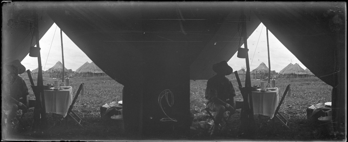 'Vy inifrån Magnus Leyers tält och ut med bord och fällstol i tältöppningen. 1 man sitter vid bordet. Toppiga hyddor och va 15 människor en bit bort. :: Enligt text till fotot: ''Looking out from M.L.´s tent''. (Tittande ut från M.L.´s tält.) ::  :: Ingår i en serie med fotonr. 5238:1-18. Se även fotonr. 5237:1-16, 5239:1-18 och 5240:1-17, 5241:1-18. ::  :: Vyer, resa till Beira, båtar, stadsmiljö. :: Nr 2.'