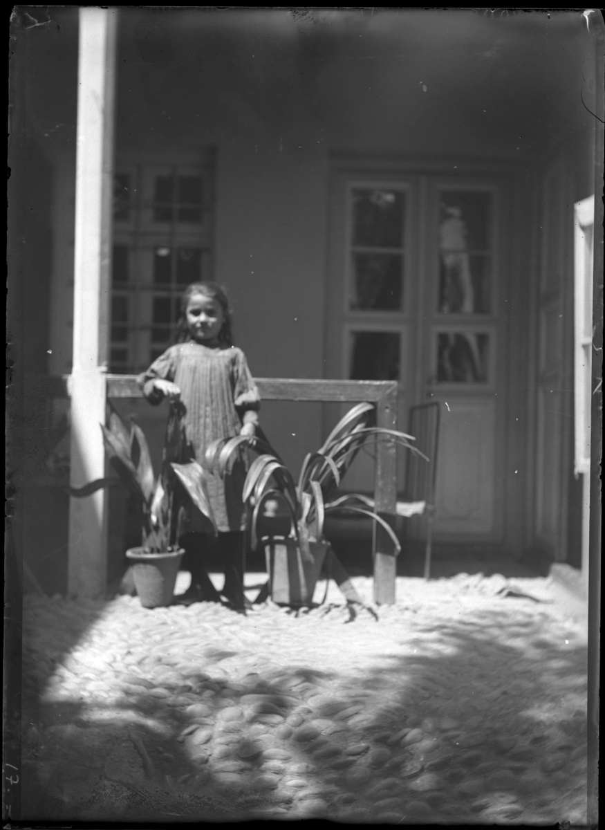 '1 flicka stående framför hus, på kullersten vid 2 blomkrukor. Kläder. Klädsel. ::  :: Ingår i serie med fotonr. 5277:1-19. Se även fotonr. 5269-5292 med bilder från Stuxbergs resa till Kaukasien.'